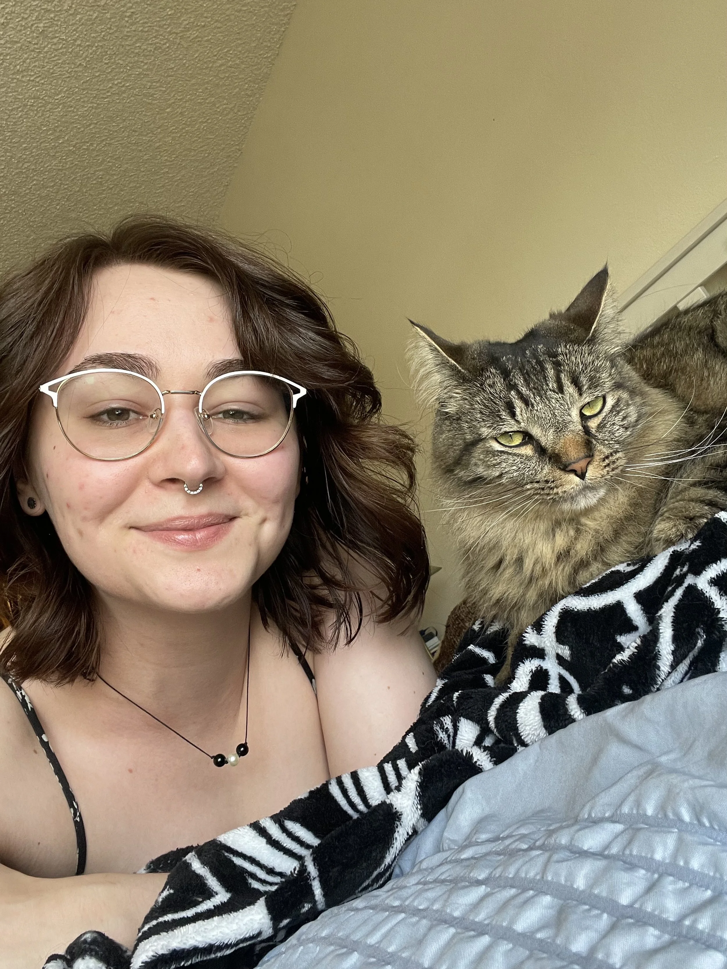 A woman with shoulder-length brown hair, glasses, and a septum piercing smiling, lying next to a tabby cat with green eyes on a bed with black and white patterned and gray bedding.