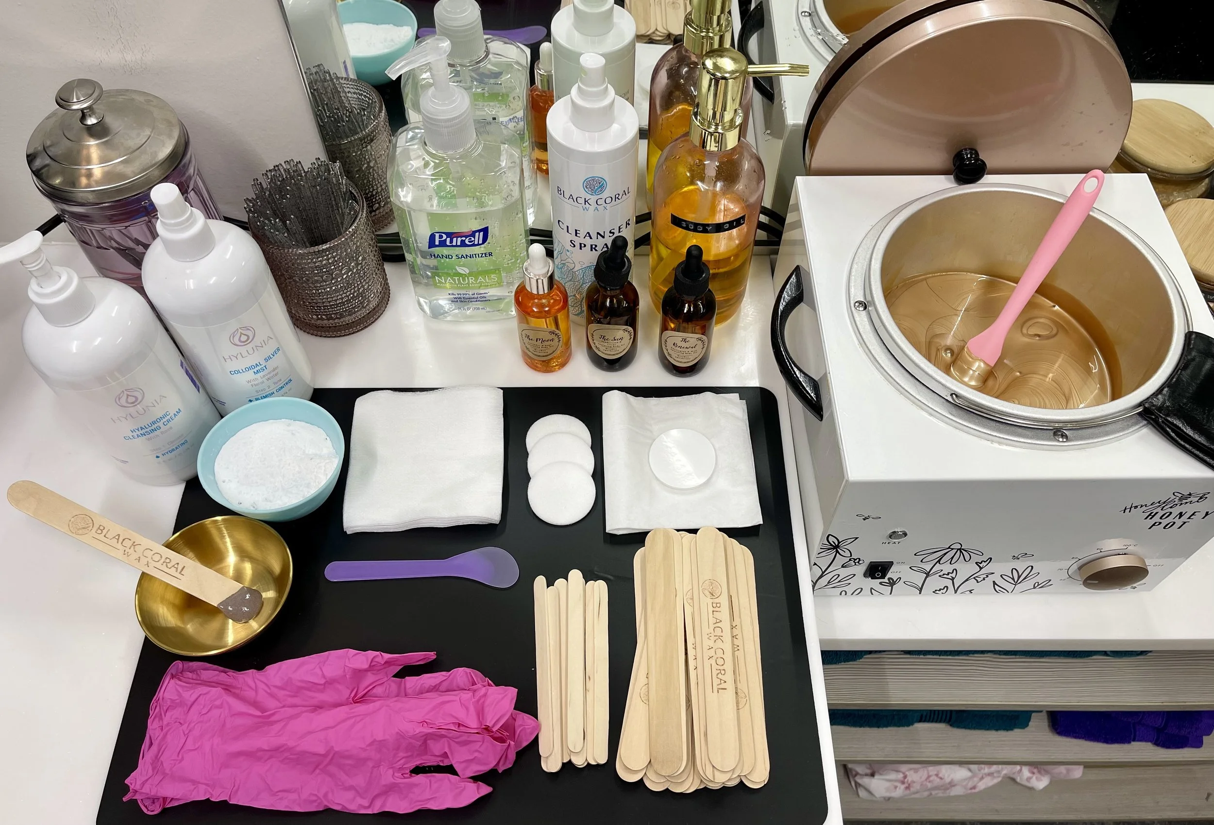 A spa or beauty treatment station with various skincare and massage products, including bottles of toner, cleanser, oils, a wax warmer with a pink spatula, wooden stir sticks, disposable gloves, cotton pads, and small containers for mixing ingredients.