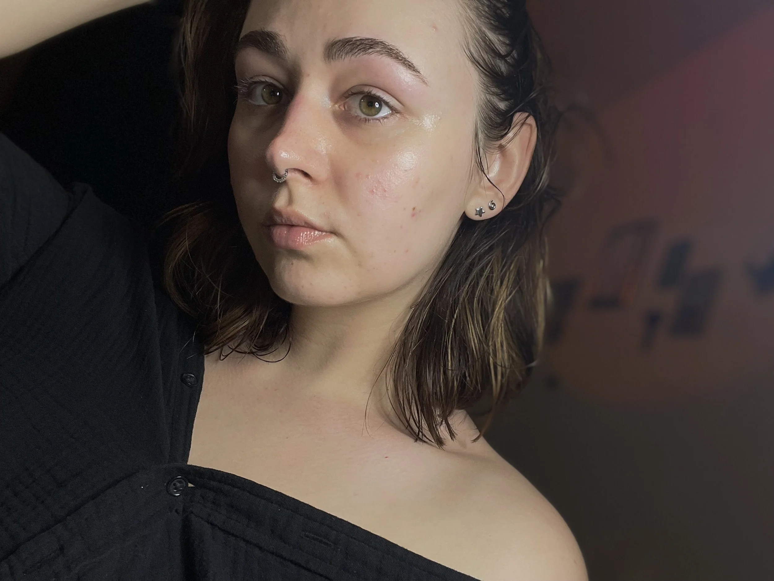 Close-up of a young woman with shoulder-length brown hair, wearing a septum piercing and multiple earrings, with a neutral expression, wearing a black top, and a blurred background.