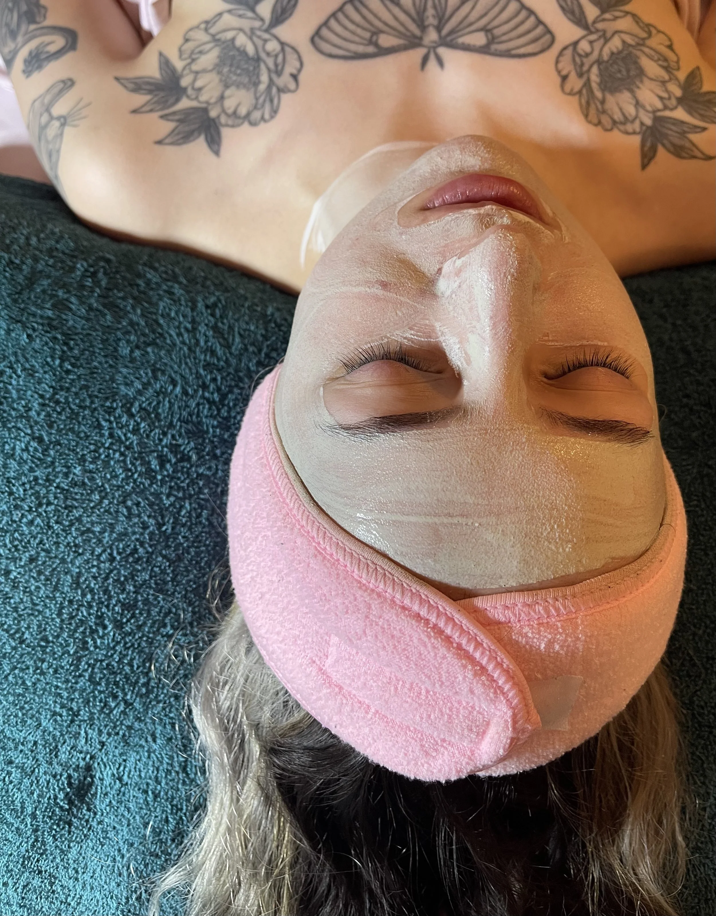 A person lying down with a facial mask, wearing a pink headband, with tattoos visible on their chest and arms, on a dark textured surface.