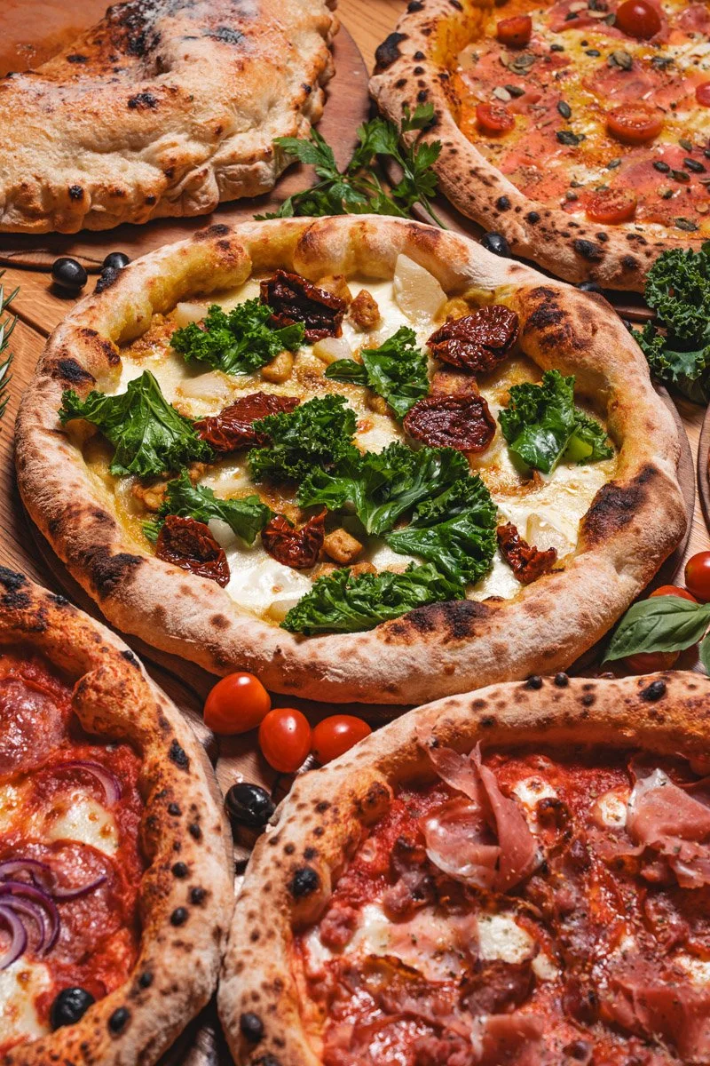 Assorted pizzas with various toppings including sun-dried tomatoes, kale, pepperoni, ham, and cherry tomatoes on a wooden table with herbs and black olives.