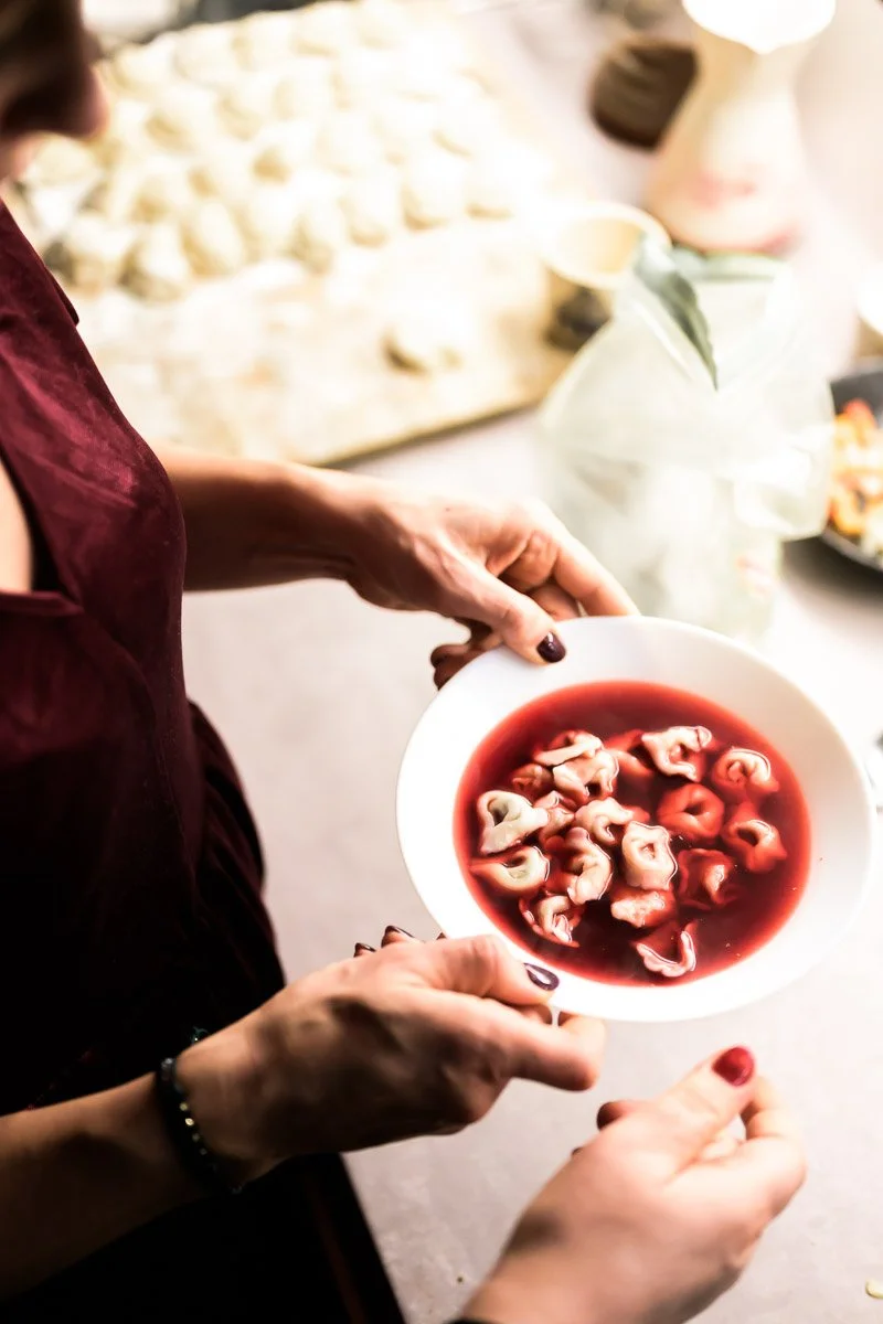 A person holding a white bowl filled with cooked tortellini in a red broth.