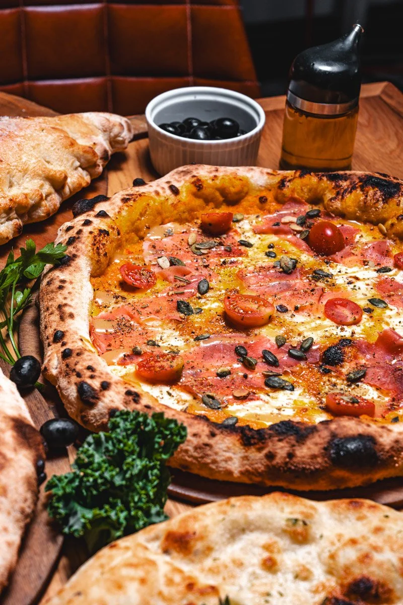 Close-up of a pizza with tomatoes, pumpkin seeds, and melted cheese on a wooden table. There are also garlic bread, a bowl of black olives, and a bottle of olive oil in the background.