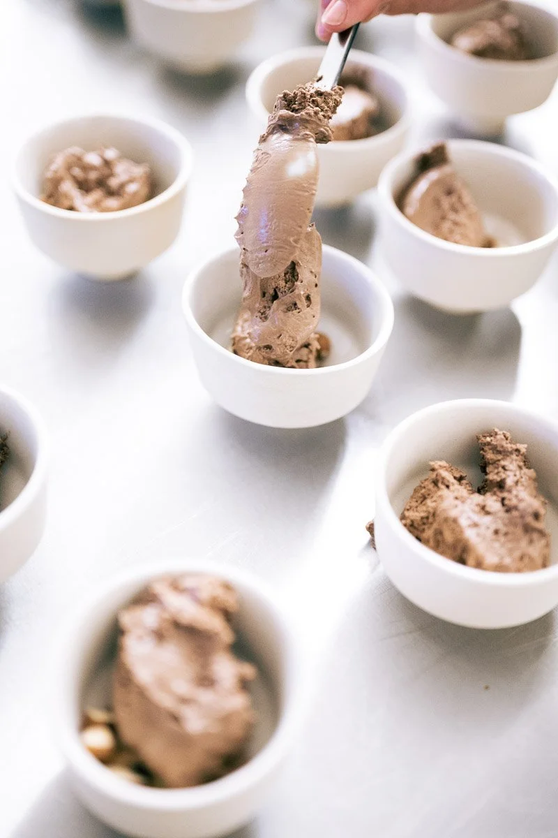 Scooping chocolate ice cream into small white bowls on a white surface.