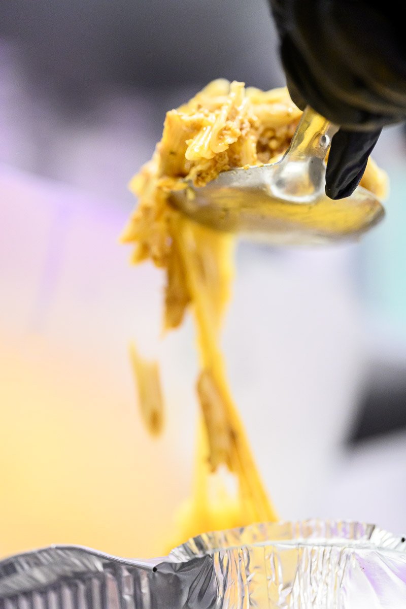 A close-up of a stainless steel ice cream scooper with a scoop of yellow confection, likely cheese or similar, dripping into an aluminum foil container.