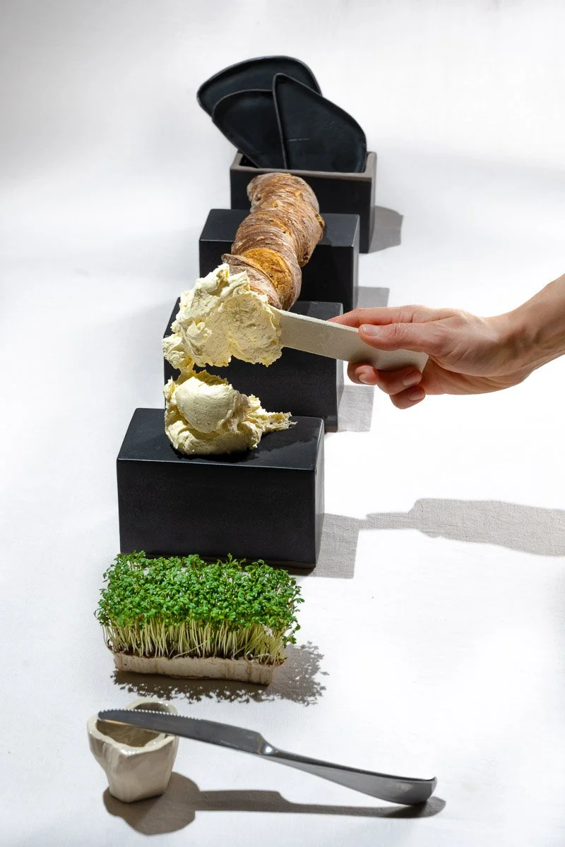A hand scooping ice cream from a large container with a root vegetable, possibly a ginger root or turmeric, on top, placed on black blocks. In the background, there are black dishes in a holder, and in the foreground, there is a container with green 