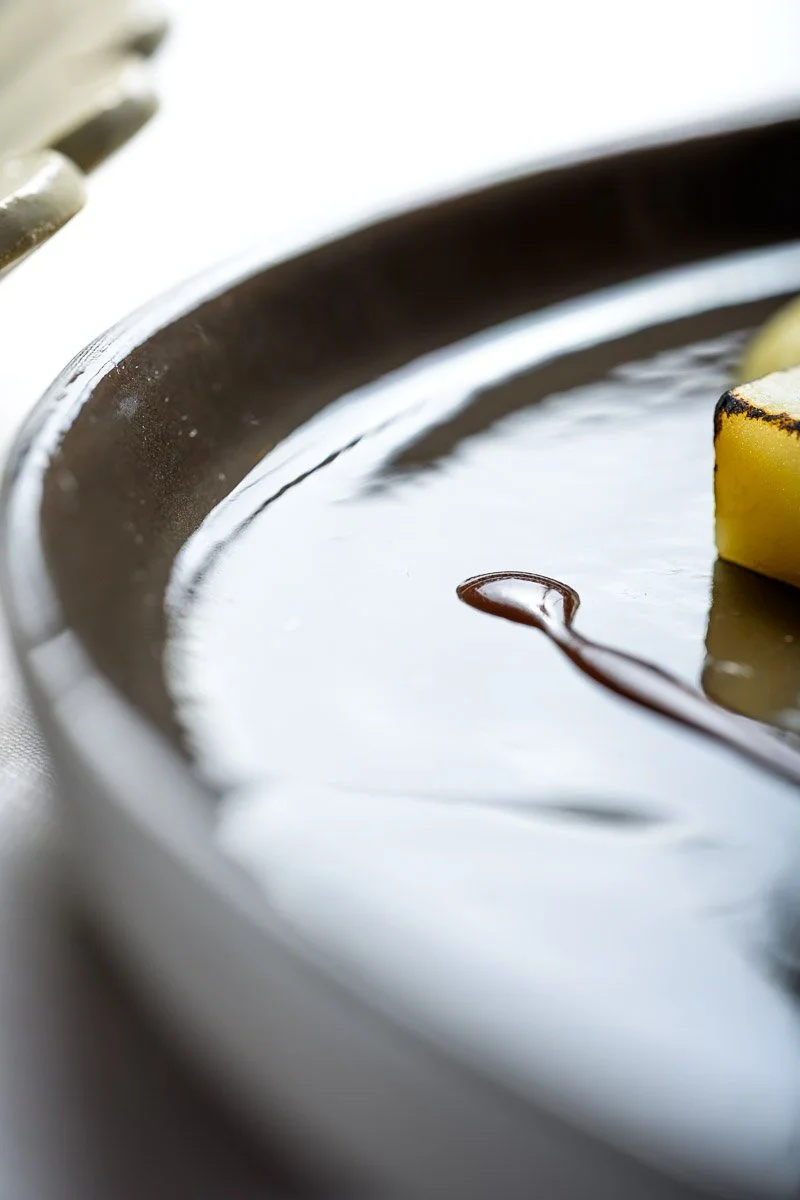 Close-up of a dark glossy surface with a small pool of chocolate syrup and a piece of cooked pineapple on the edge.