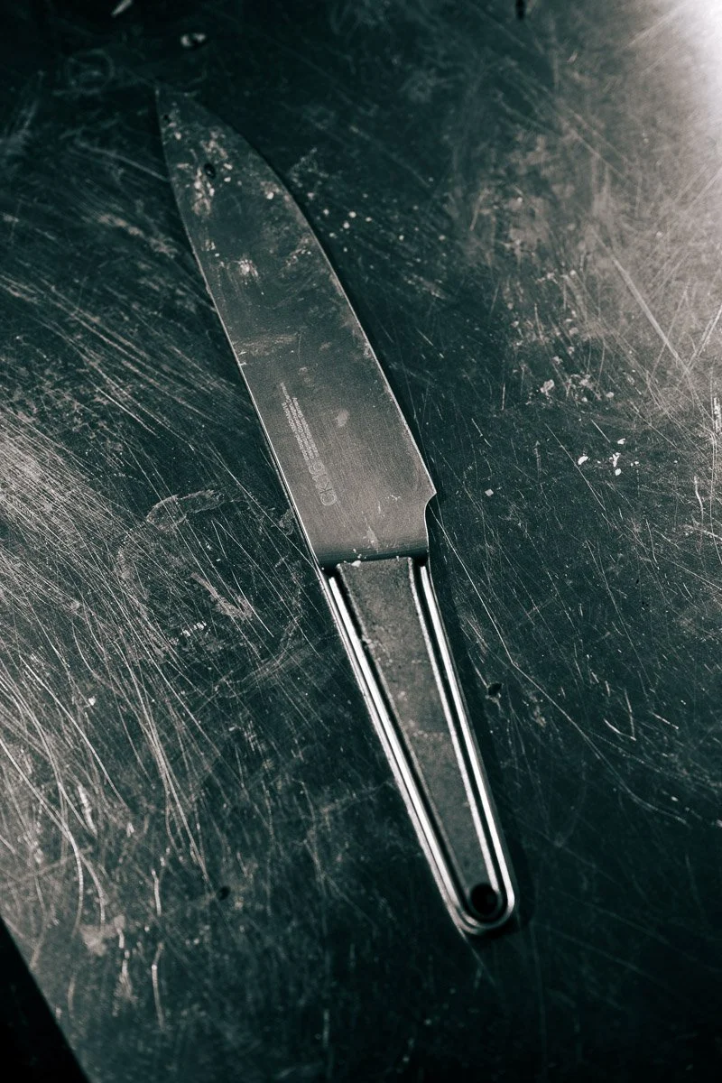 A large kitchen knife with a black handle resting on a scratched, metallic surface.
