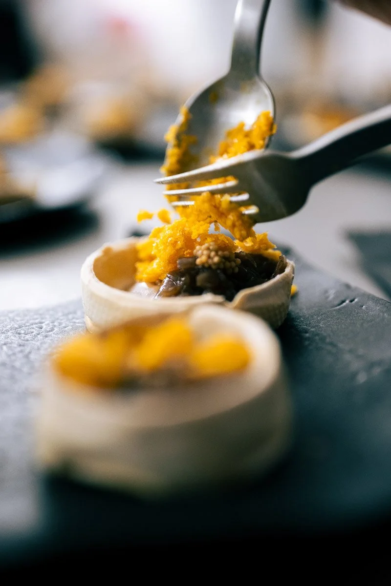 Close-up of a deviled egg topped with yellow and dark seasoning, with a silver fork and spoon above it, on a dark surface.