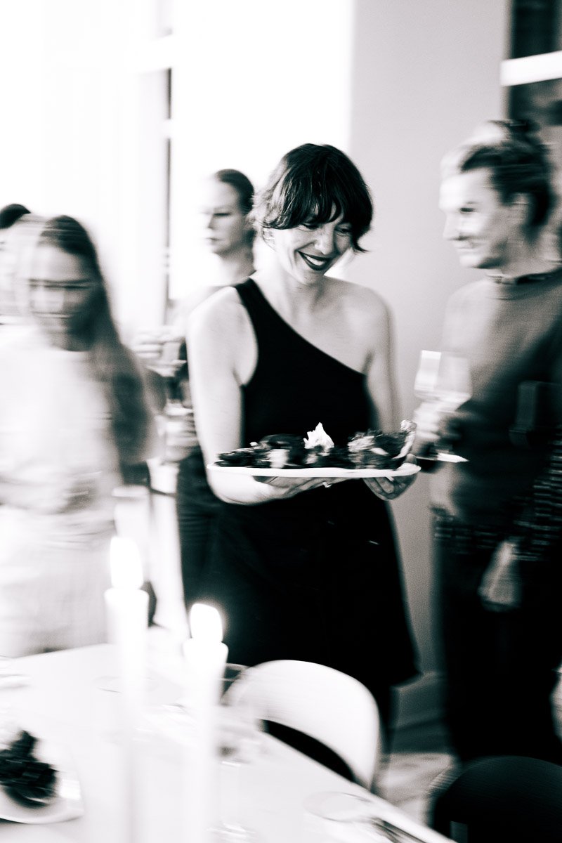 A woman in a black dress holding a tray of desserts at a party or gathering, with other blurred people in the background.