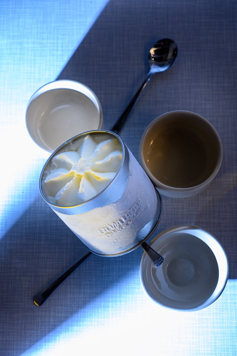 A glass of yogurt topped with whipped cream, four empty cups, and a spoon on a table with a light blue surface.