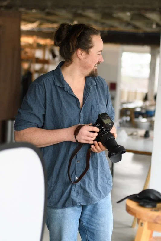 A man with long hair in a bun, wearing a blue button-up shirt and jeans, holding a camera, and smiling while looking to the side inside a rustic, well-lit space.