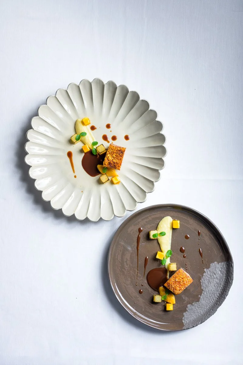 Two plates with modern dessert presentations featuring small cake cubes, green garnishes, banana slices, and drizzles of sauce, on a white surface.