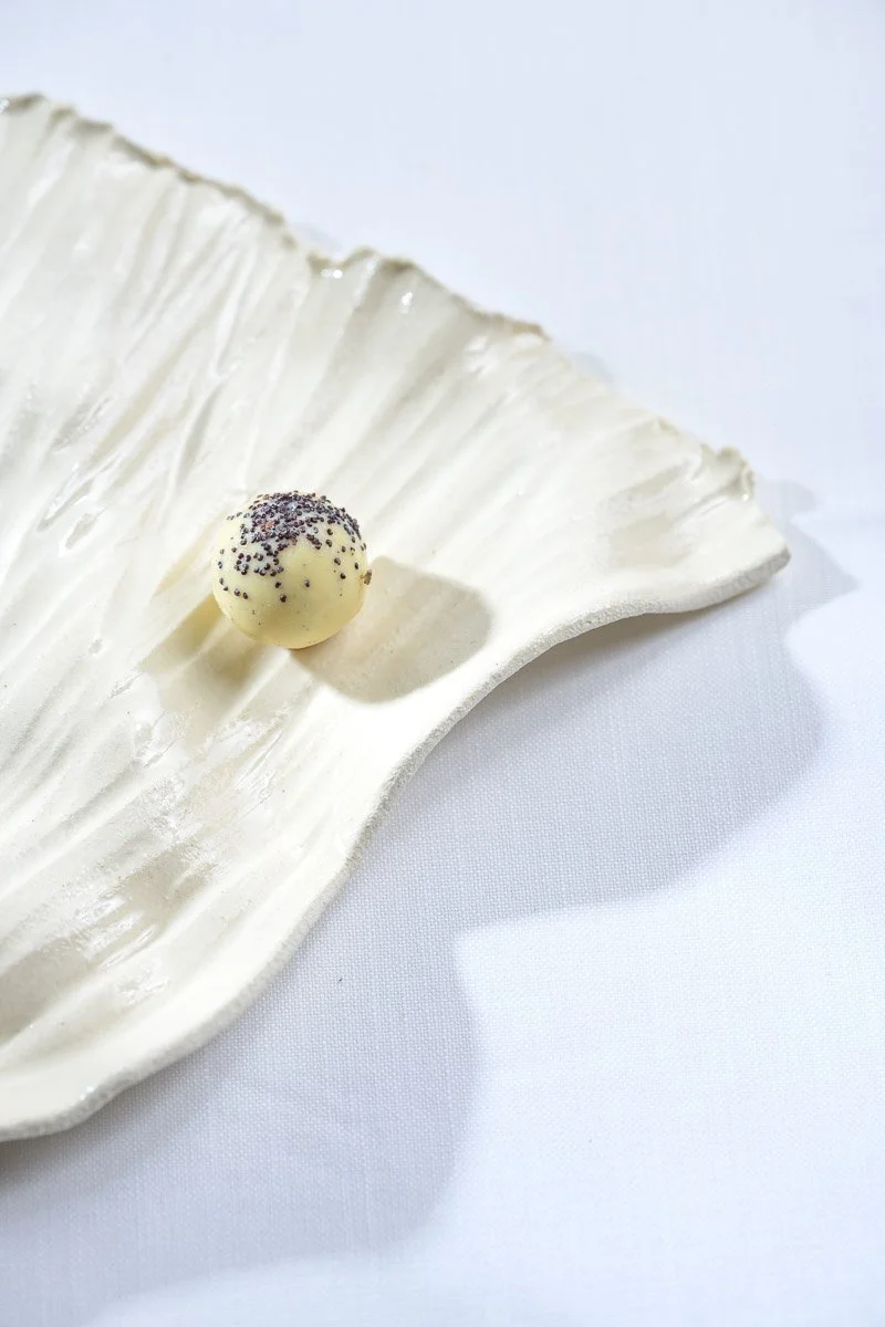 A white chocolate truffle with black sprinkles on top, resting on a decorative white ceramic leaf plate with embossed vein details, against a white background.