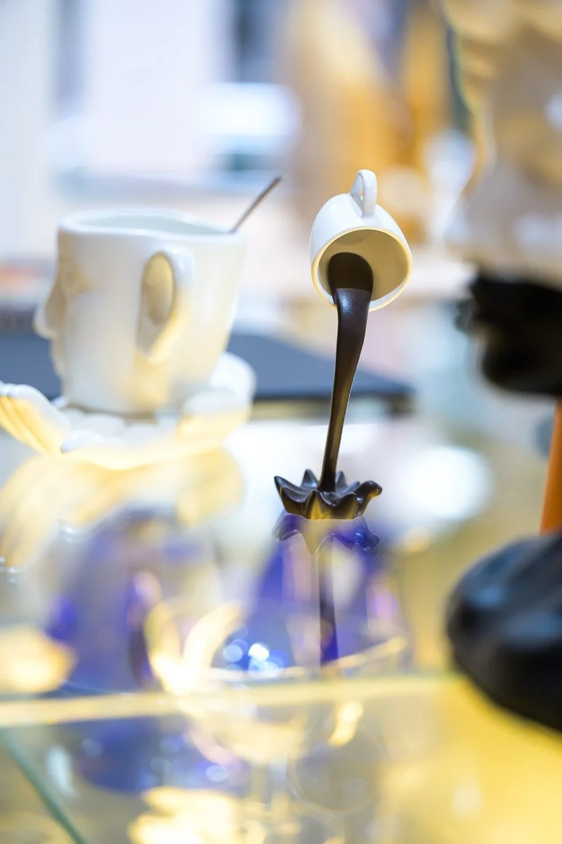 A white mug with a face design and a spoon inside, a teapot shaped like a bird pouring chocolate, and a blurred background with other objects and a yellowish color palette.