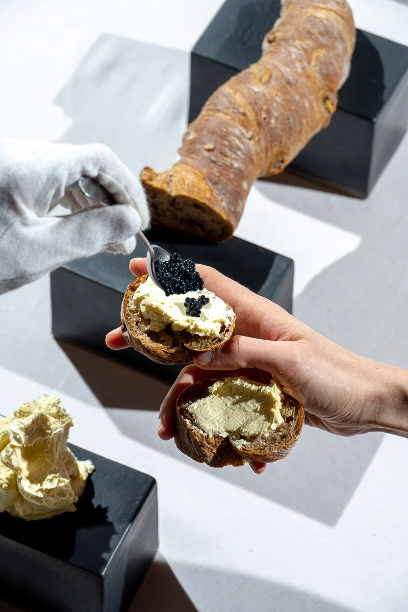 Person spreading butter on a bread roll with black caviar on top, while another bread roll with butter is held nearby. A large yam and a dollop of butter are on black display blocks.