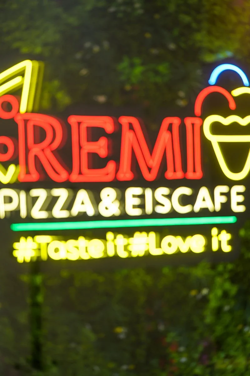 Neon sign at a pizza and ice cafe with the words 'Remi Pizza & Eiscafe', a colorful ice cream cone graphic, and hashtags '#TasteIt #LoveIt'