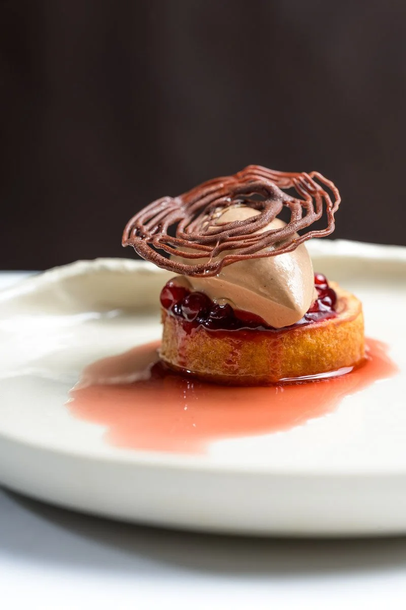 A plated dessert featuring a round pastry topped with red berries, a dollop of cream or mousse, and decorative chocolate garnish on a white plate.