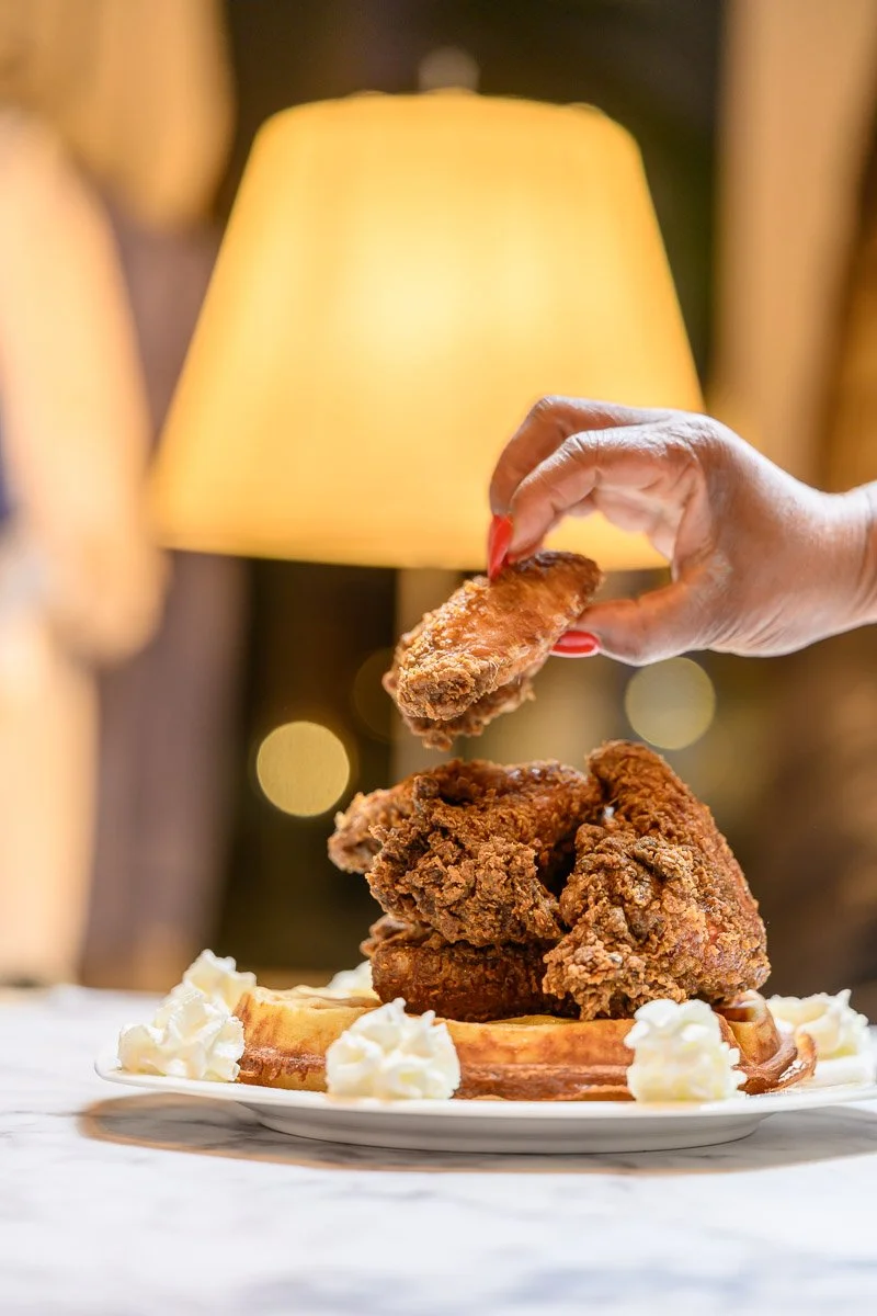 A person with red-painted nails is dropping a piece of fried chicken onto a plate with more pieces of fried chicken and waffles topped with whipped cream, in front of a yellow lampshade.