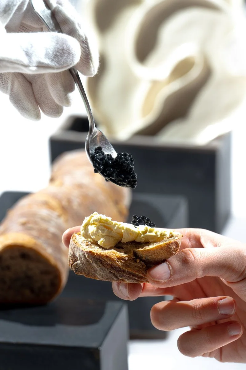 Person holding bread with butter and black caviar being added, with a breadstick in the background.