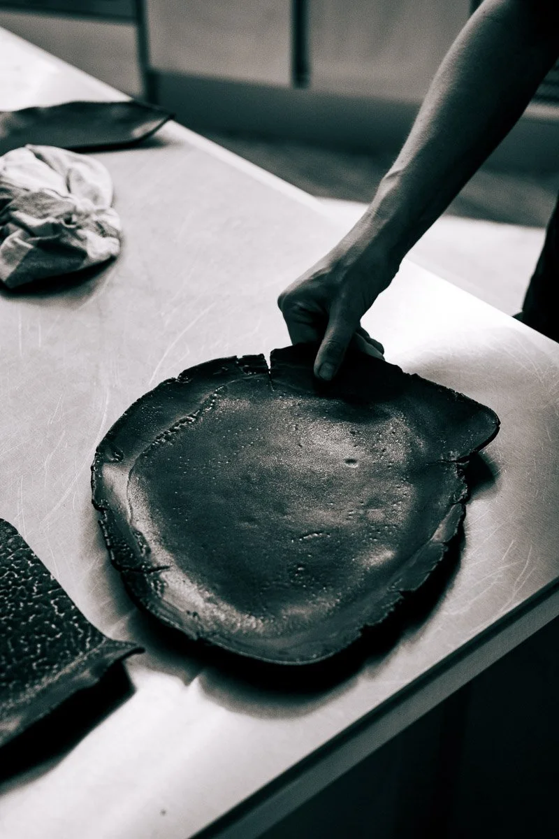A person is handling a large, melted piece of charcoal or black material on a stainless steel surface in a kitchen or workshop setting.