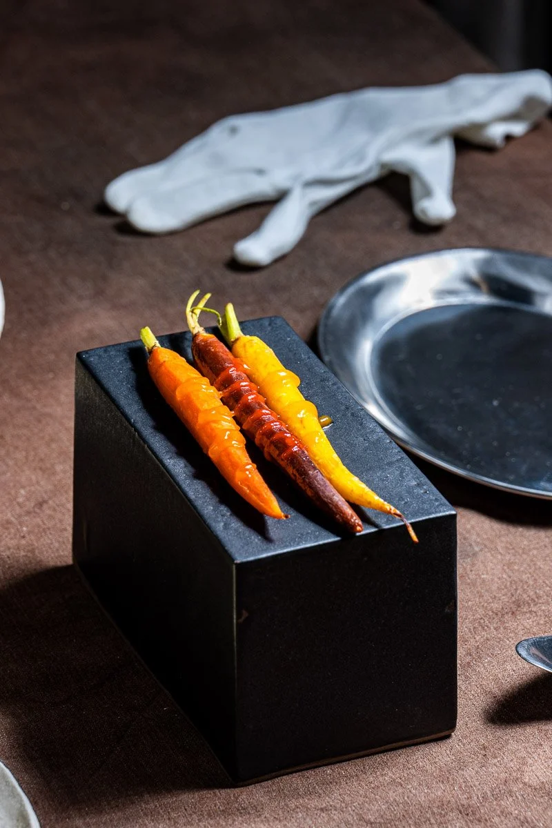 Three colorful carrots — orange, purple, and yellow — are placed on a black rectangular stand on a brown table. There is a shiny silver plate and a white cloth or napkin in the background.