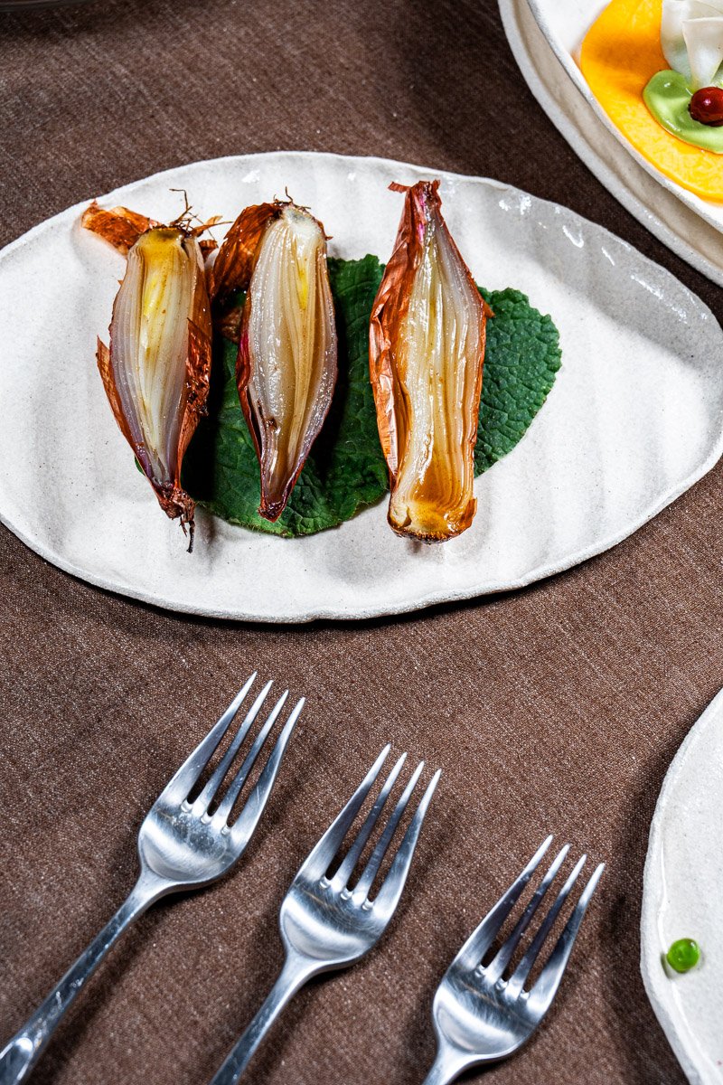 Three roasted shallots on a white oval plate garnished with a green leafy herb, placed on a brown table.