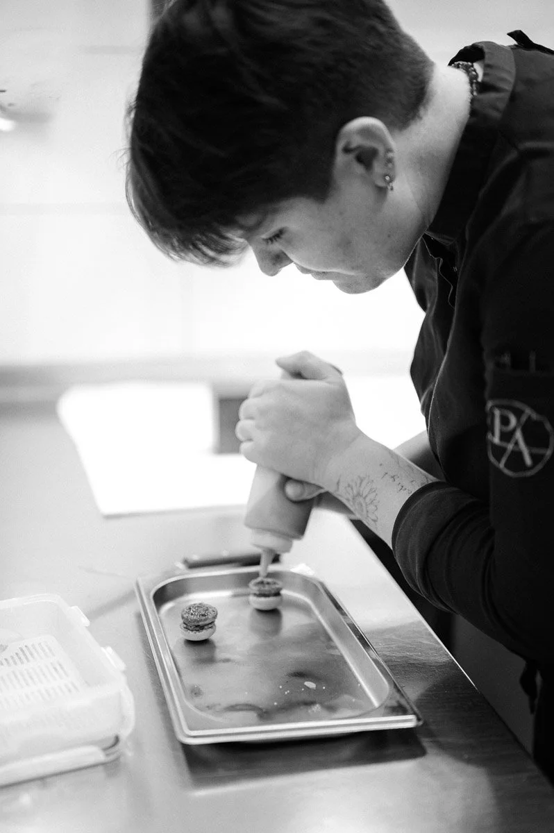 A person with short hair and earrings decorates a cupcake with frosting in a professional kitchen.