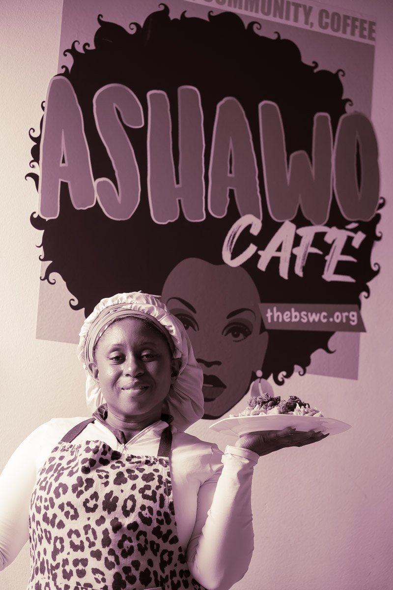 A woman dressed as a chef wearing a leopard print apron and a chef's hat, holding a plate of food in front of a sign that reads 'ASHAWO CAFE' with a stylized illustration of a woman's face with curly hair.