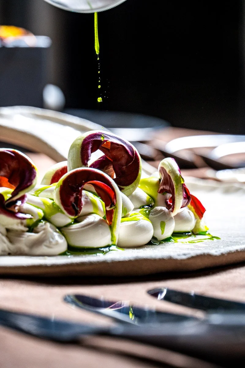 A plate of burrata cheese topped with radicchio and drizzled with olive oil, with droplets falling from a spoon or utensil above.