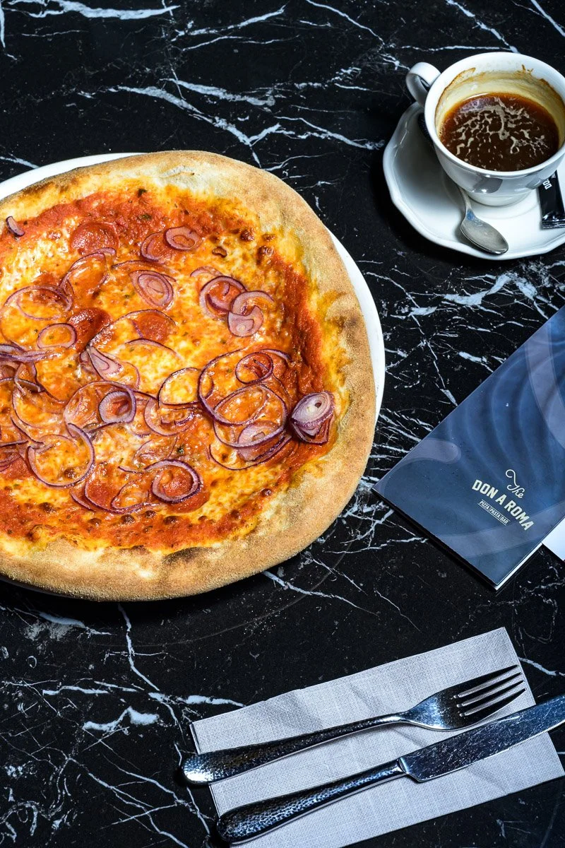 A pizza with cheese and sliced red onions on a white plate, a cup of coffee on a saucer with a spoon, a fork and knife on a napkin, and a menu on a black marble table.