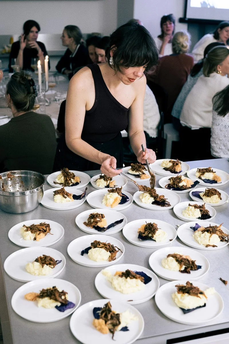 A woman in a black tank top is serving food onto white plates at a banquet or event, with a group of people seated at tables in the background.
