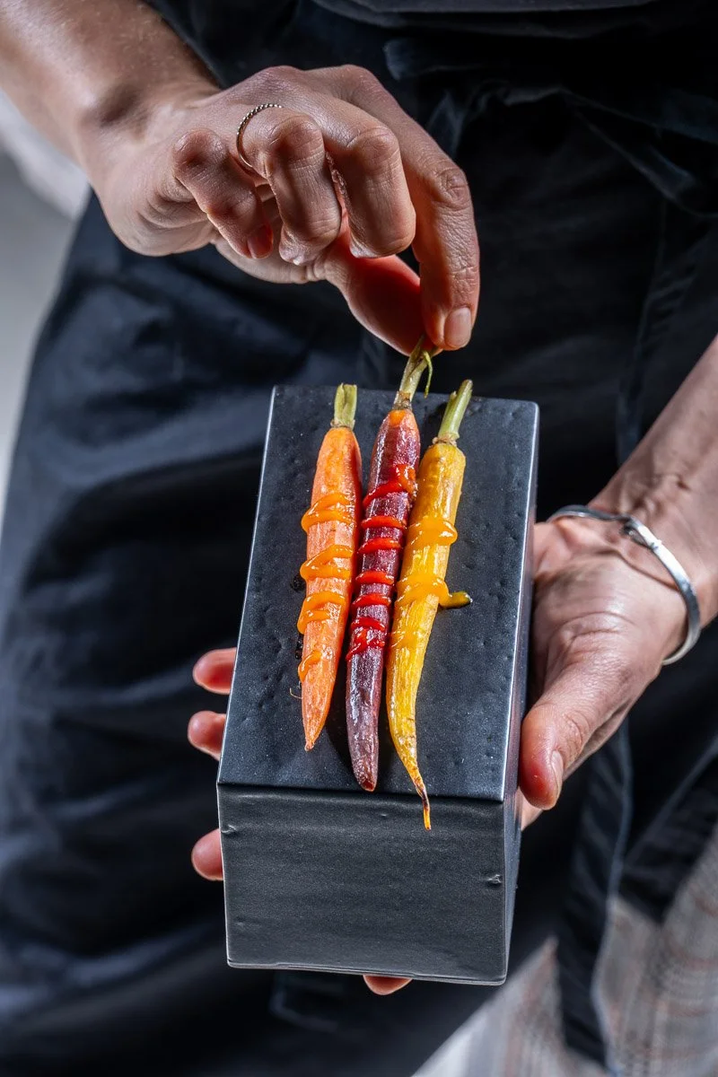 A person holding a small black rectangular plate with three colorful, carrot-shaped pastries decorated with different colored sauces on top.