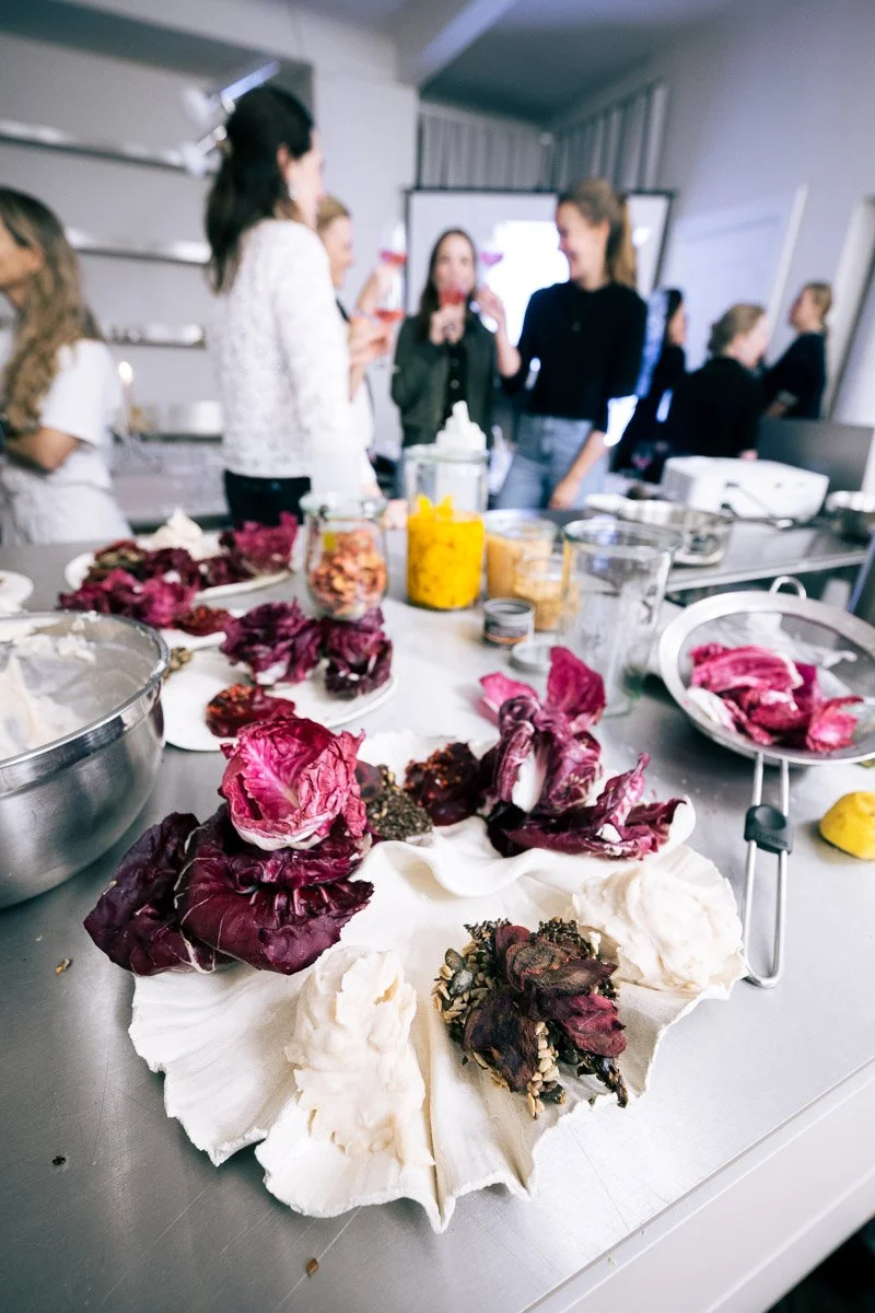 A table with flower petals and herbs, and a group of women in the background during a workshop or social gathering.