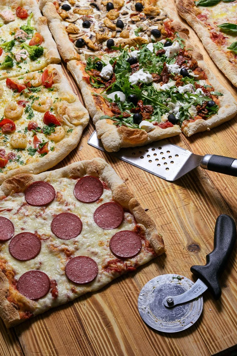 Four different types of rectangular pizzas on a wooden pizza peel. The pizzas include a pepperoni pizza, a veggie pizza with cherry tomatoes and green herbs, a chicken and black olive pizza, and a prosciutto and arugula pizza with drizzled cheese.