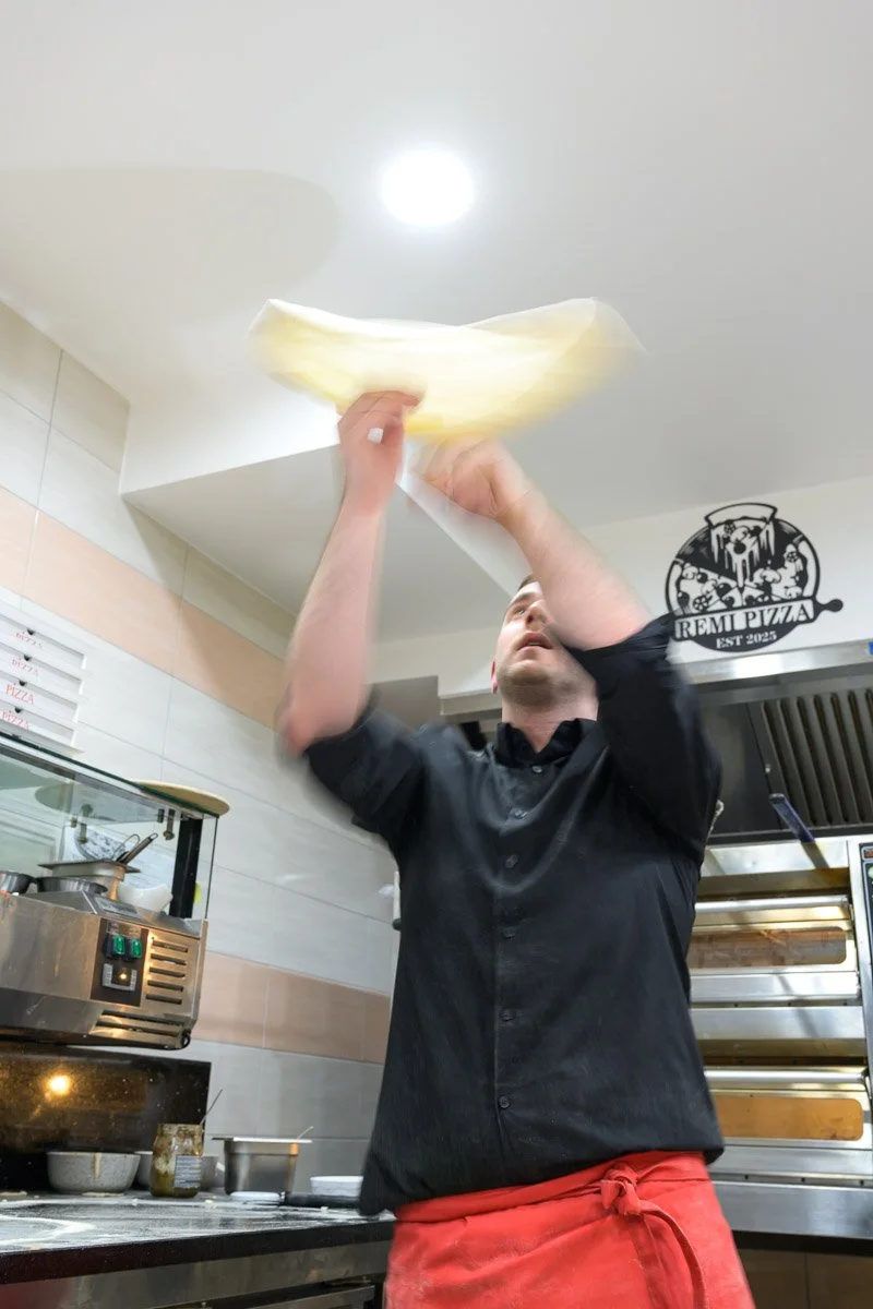 A person tossing pizza dough in the air inside a pizzeria kitchen.