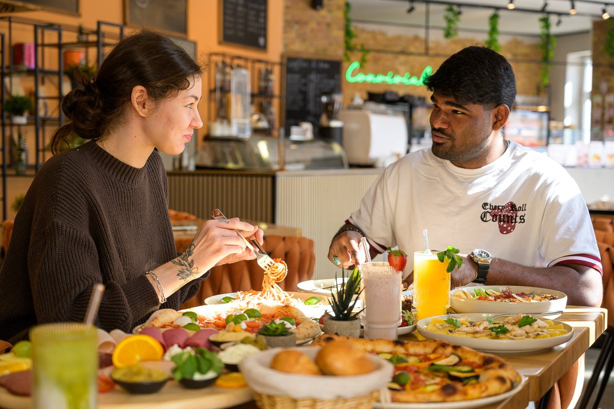 A woman and a man sit across from each other enjoying a meal at a restaurant, with various dishes including pizza, pasta, and drinks on the table.