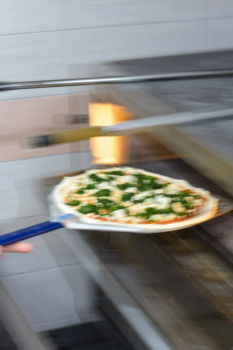 A pizza with green leafy vegetables being placed in an oven with a pizza peel.