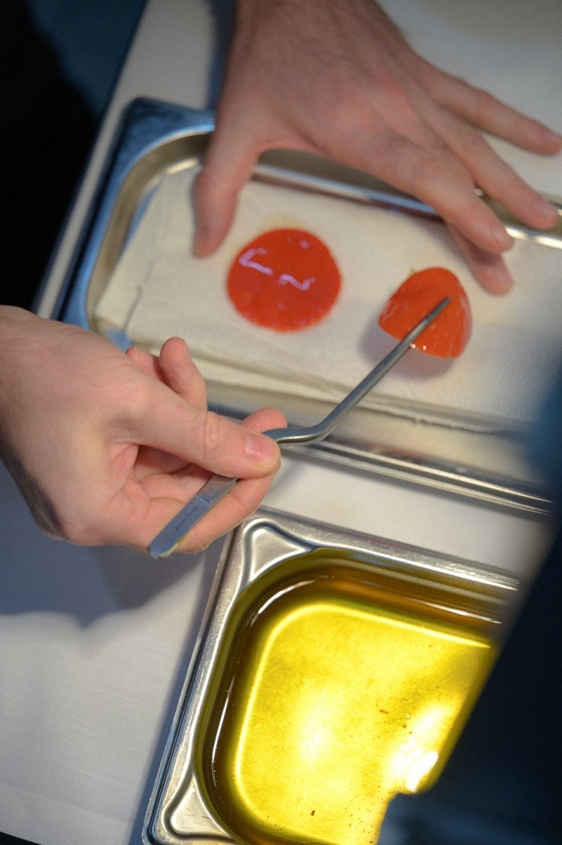 Person preparing or handling two orange spheres, possibly bait or decorative objects, on a metal tray with a pool of greenish-yellow liquid nearby.