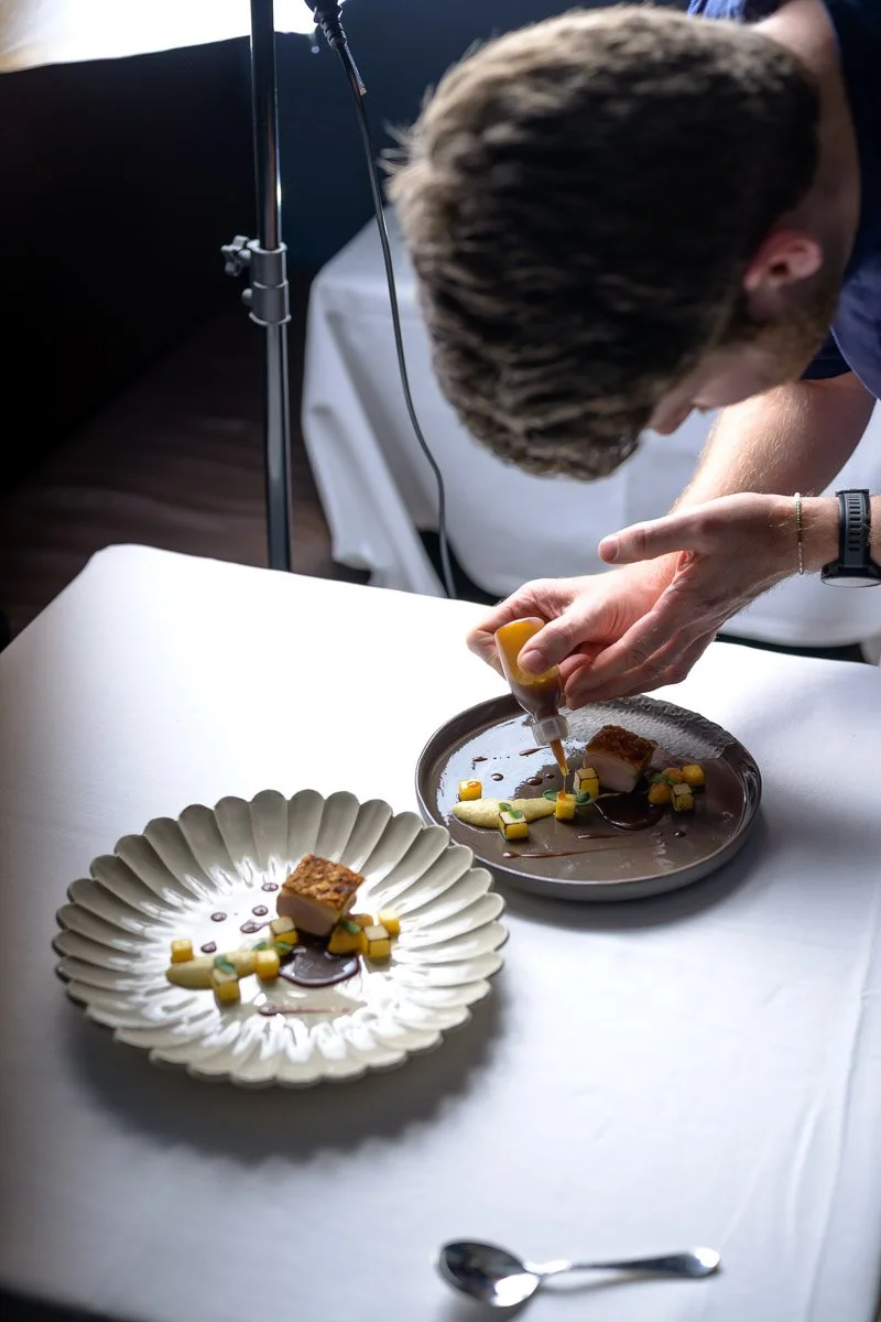 A chef or food stylist is carefully plating a gourmet dish with a squeeze bottle, adding sauce to a plate that has a piece of meat, vegetables, and sauce artfully arranged. There is another plate with a similar presentation on the table, and professi