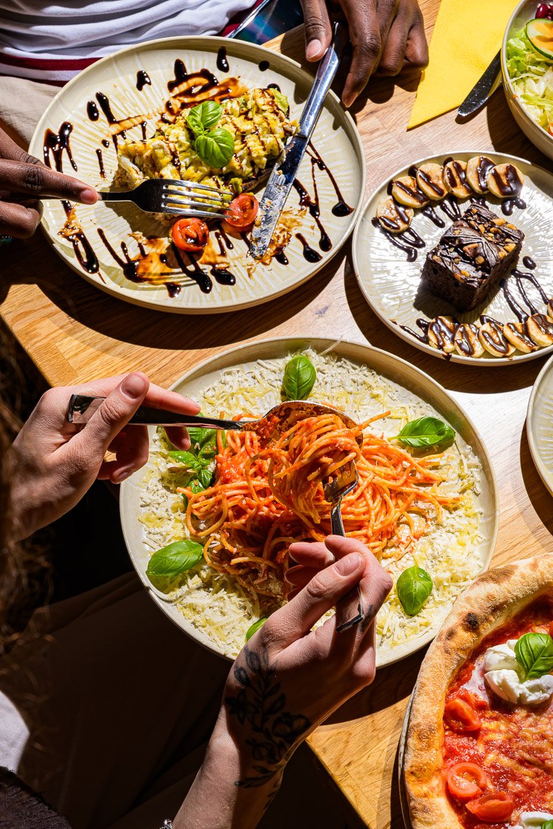 Several dishes on a wooden table including a plate of spaghetti with basil, a salad, a slice of chocolate cake with caramel and nuts, a pizza with tomato, mozzarella, basil, and cherry tomatoes, and a salad with lettuce and cucumbers. Two people are 