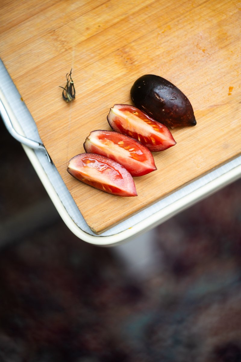 A wooden cutting board on a table with sliced tomato halves and an uncut tomato, along with a small insect on the side.