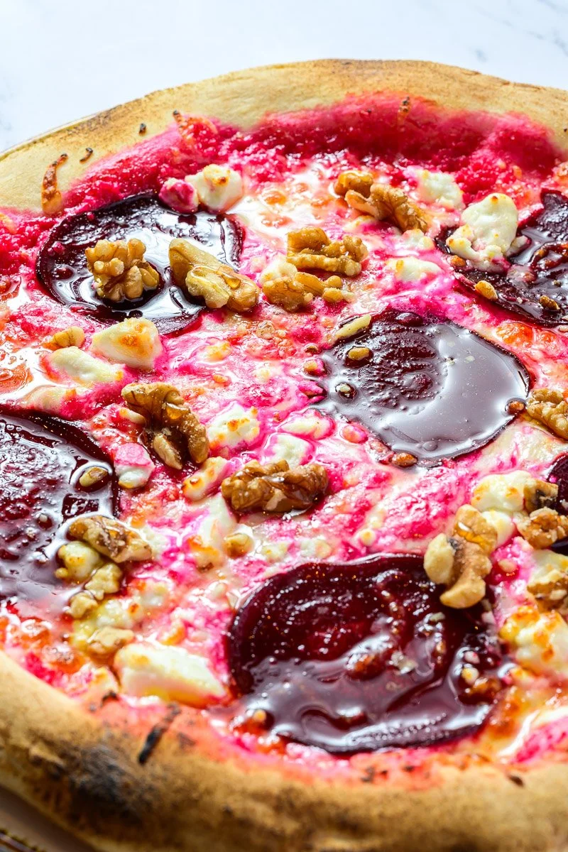 Close-up of a pizza with a golden crust, red tomato sauce, white cheese, black olive slices, and sprinkled walnuts.