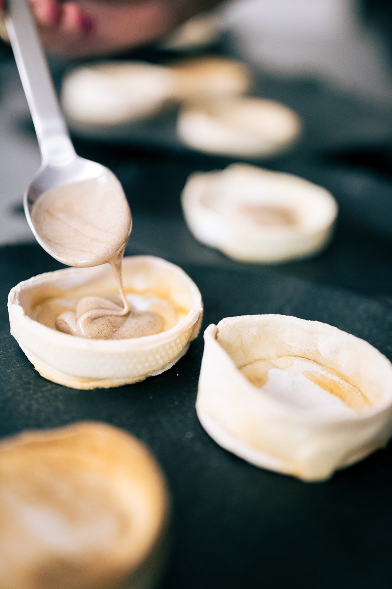 A spoon with chocolate filling being added to tart shells on a dark surface.