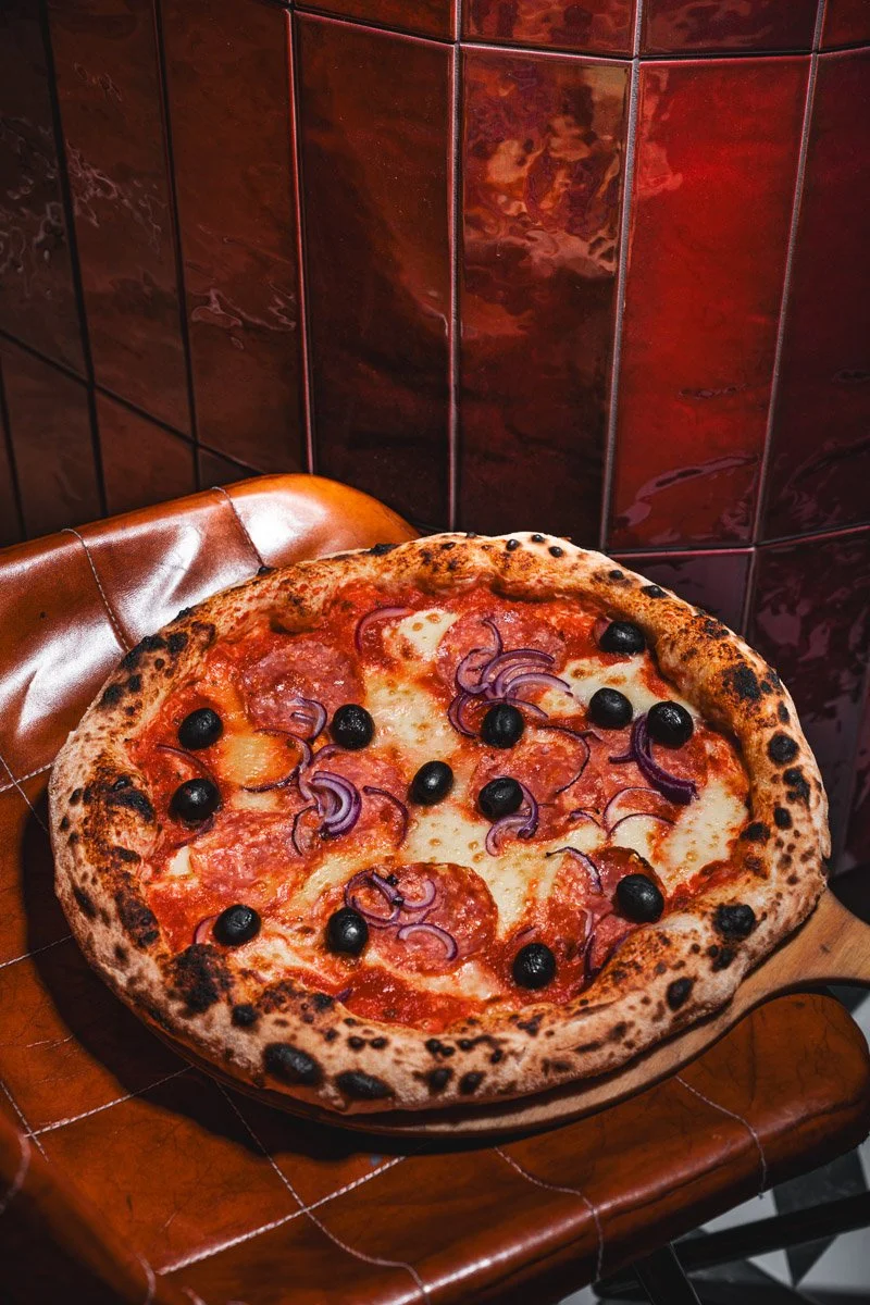 A pizza with black olives, sliced red onions, pepperoni, and cheese on a wooden pizza peel, placed on a leather chair in a restaurant with dark tiled walls.