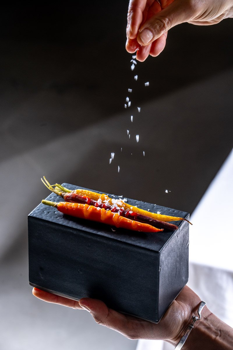 A hand sprinkling salt onto colorful carrots placed on a black box.
