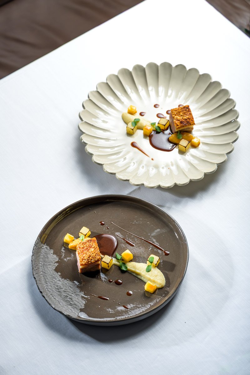 Two plates with small, artfully arranged portions of gourmet food, including a cube of crispy textured protein or cake, small green herbs, yellow diced vegetables, a smear of white sauce, and drizzle of brown sauce, on a white tablecloth.