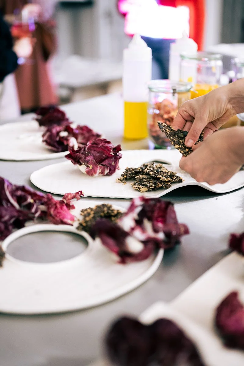 Person preparing or assembling edible seed or nut snack bars with seeds, nuts, and honey on a white plate surrounded by dried flower petals on a table.