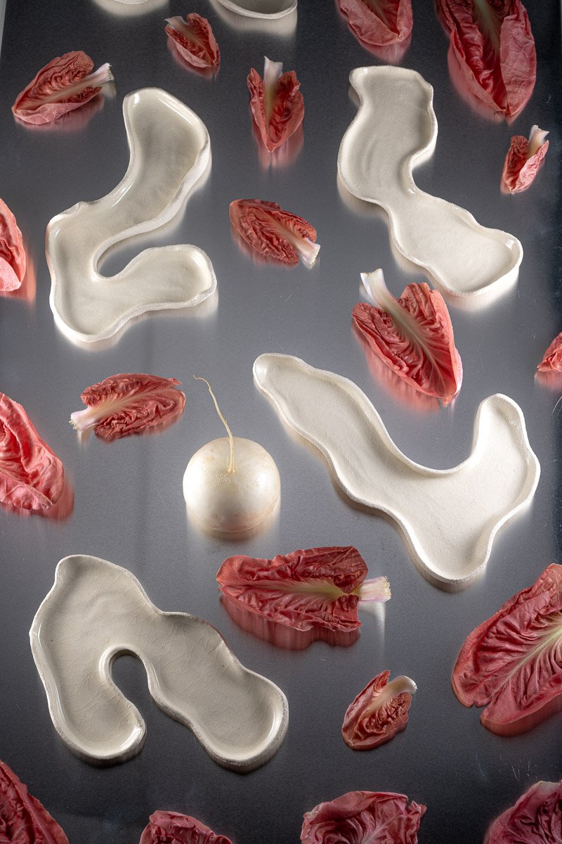 A white radish with a green stem, surrounded by sliced red radicchio leaves and various white ceramic dish pieces on a reflective gray surface.