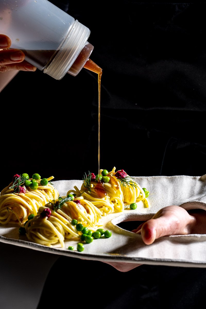 Person pouring sauce over plated pasta garnished with green peas and herbs, against a dark background.